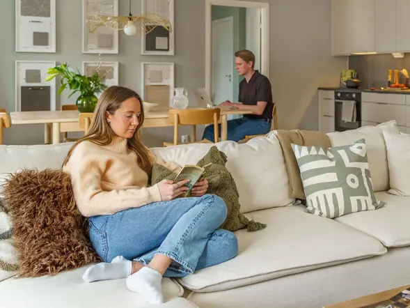 Person sitter i soffa medan en annan sitter vid köksbordet i en modern bostad.