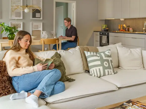 Person sitter i soffa medan en annan sitter vid köksbordet i en modern bostad.